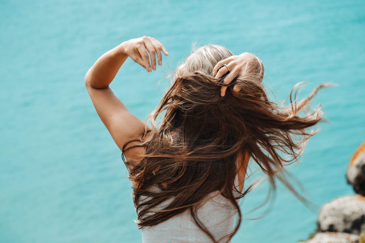 Photo by Helena I Back view of a woman with flowing hair near the sea in Portugal, capturing a moment of solitude.