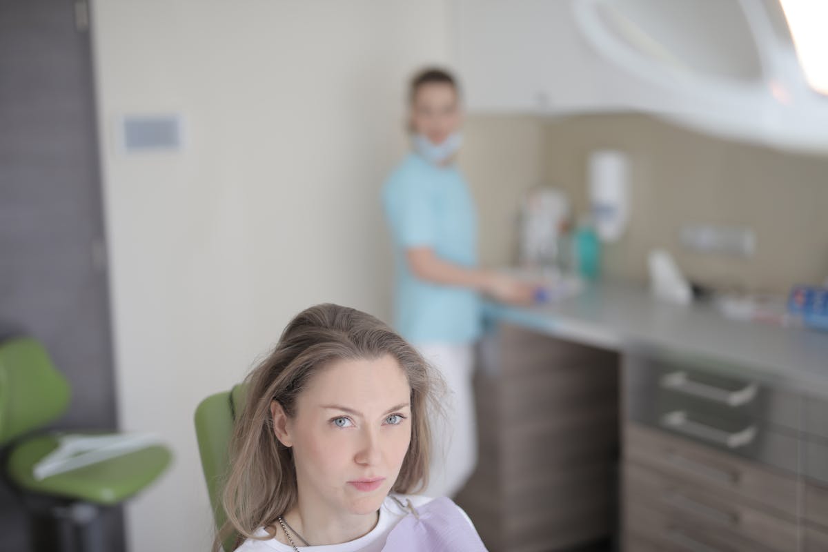 Woman in dental clinic with dentist preparing in background, modern healthcare setting.