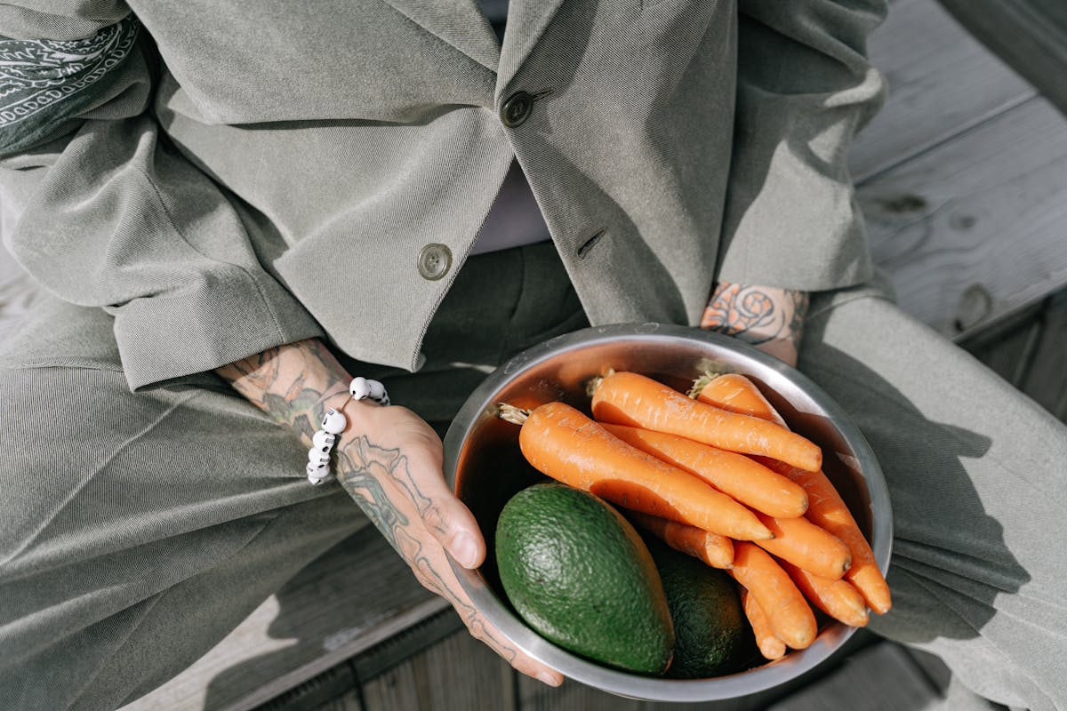 Photo by Ivan S Person in gray suit holding a bowl of fresh carrots and avocados outdoors.