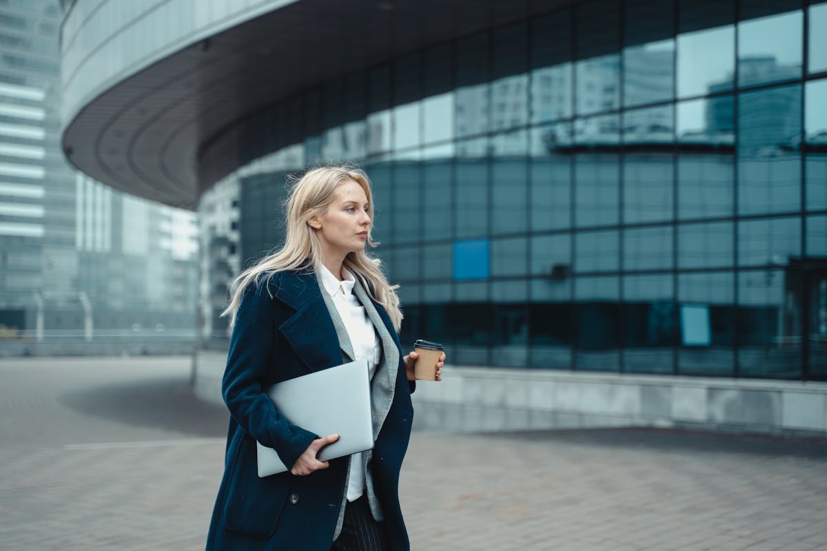 Confident businesswoman walking outside modern office building holding laptop and coffee.