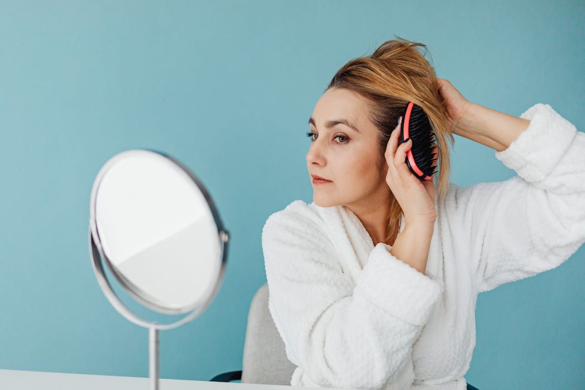 Caucasian woman wearing bathrobe and brushing hair while looking in mirror indoors.