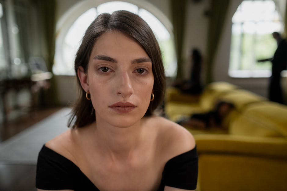 Portret młodej kobiety o naturalnym makijażu, siedzącej w eleganckim wnętrzu z żółtą sofą w tle. Kobieta ma krótkie, brązowe włosy i nosi czarną bluzkę bez rękawów. W tle widać rozmyte sylwetki innych osób.